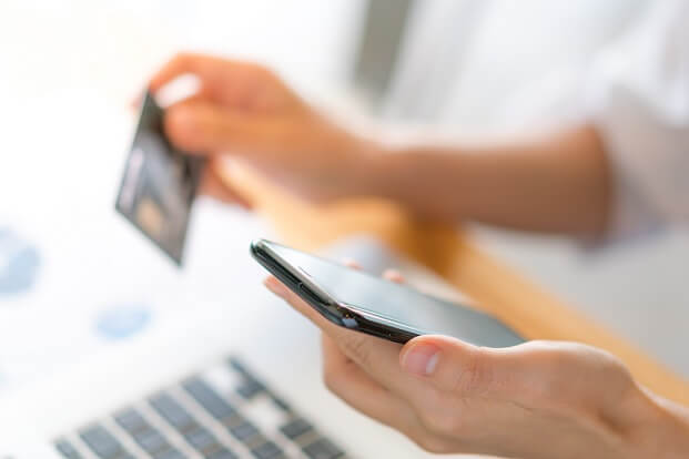 mãos femininas seguram um smartphone e um cartão de crédito em frente a um notebook, dando a ideia de compras em loja virtual
