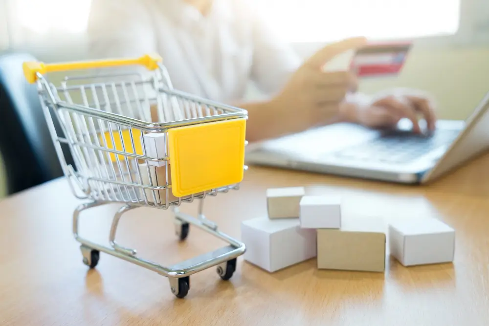 mini Carrinho de compras em cima de mesa com pessoa segurando cartão de crédito desfocada no fundo
