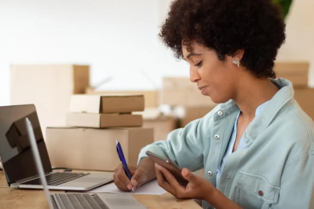 mesa com 2 laptops, mulher segurando um celular enquanto anota em papel com caneta