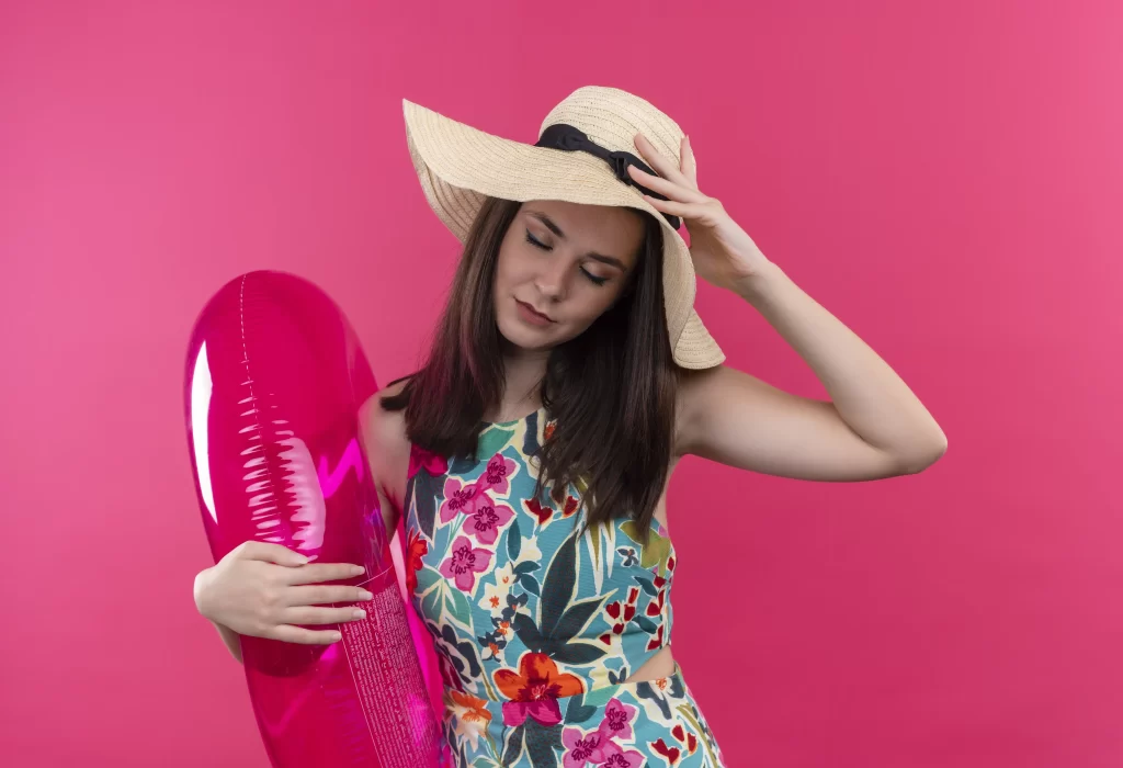 mulher vestindo maiô, posando para foto segurando uma boia e usando chapéu de praia
