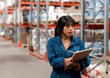 Mulher realizando inventário de estoque em depósito, anotando dados em prancheta entre prateleiras com produtos armazenados.