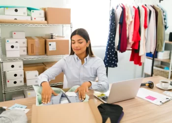 Lojista organizando estoque com peças recebidas de fornecedores de roupas, em ambiente de loja virtual com araras e caixas para envio