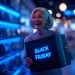 Consumidora sorridente segura uma caixa com o letreiro “Black Friday” em luz neon azul dentro de uma loja iluminada, representando o clima de compras e promoções da Black Friday 2025.