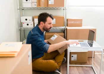 Homem sentado no chão de estoque utilizando laptop segurando caixa de envio em uma das mãos.