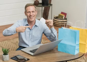 Homem branco segurando um cartão sentado à mesa com laptop e sacolas de compras