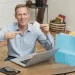 Homem branco segurando um cartão sentado à mesa com laptop e sacolas de compras